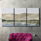 Three-panel canvas wall art featuring desert mountain landscape watercolor, displayed in a living room above a suede accent chair.