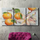 Three-panel canvas wall art featuring bright tangerine still life blossoms, displayed in a living room above a suede accent chair.