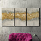 Three-panel canvas wall art featuring flowing abstract golden tones, displayed in a living room above a suede accent chair.