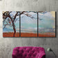 Three-panel canvas wall art featuring coastal trees at evening light, displayed in a living room above a suede accent chair.
