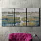Three-panel canvas wall art featuring pastoral marshland landscape, displayed in a living room above a suede accent chair.