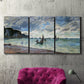 Three-panel canvas wall art featuring figures walking along sandy shoreline, displayed in a living room above a suede accent chair.