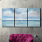 Three-panel canvas wall art featuring tropical clouds over ocean horizon, displayed in a living room above a suede accent chair.