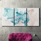Three-panel canvas wall art featuring soft abstract watercolor forms, displayed in a living room above a suede accent chair.