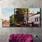 Three-panel canvas wall art featuring riverside houses along canal, displayed in a living room above a suede accent chair.