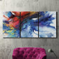 Three-panel canvas wall art featuring vibrant abstract color explosion, displayed in a living room above a suede accent chair.