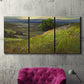 Three-panel canvas wall art featuring winding path through rolling hills, displayed in a living room above a suede accent chair.