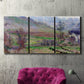 Three-panel canvas wall art featuring cliffside cottage overlooking sea, displayed in a living room above a suede accent chair.