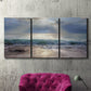 Three-panel canvas wall art featuring soft sunrise over open water, displayed in a living room above a suede accent chair.
