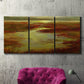 Three-panel canvas wall art featuring coastal shoreline with warm light, displayed in a living room above a suede accent chair.