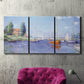 Three-panel canvas wall art featuring river flowing past riverside town, displayed in a living room above a suede accent chair.