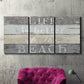 Three-panel canvas wall art featuring beach themed typography design, displayed in a living room above a suede accent chair.