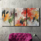 Three-panel canvas wall art featuring abstract floral color composition, displayed in a living room above a suede accent chair.