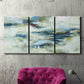 Three-panel canvas wall art featuring dreamlike abstract drifting motion, displayed in a living room above a suede accent chair.