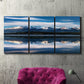 Three-panel canvas wall art featuring calm lake beneath mountains photograph, displayed in a living room above a suede accent chair.