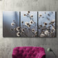 Three-panel canvas wall art featuring delicate spring blossoms on branches, displayed in a living room above a suede accent chair.