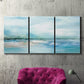 Three-panel canvas wall art featuring sunset silhouette over coastal water, displayed in a living room above a suede accent chair.