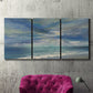 Three-panel canvas wall art featuring sunbeams breaking through coastal clouds, displayed in a living room above a suede accent chair.