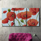 Three-panel canvas wall art featuring red poppies in motion, displayed in a living room above a suede accent chair.