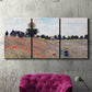 Three-panel canvas wall art featuring poppy field stretching across meadow, displayed in a living room above a suede accent chair.