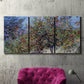 Three-panel canvas wall art featuring orchard of trees in spring bloom, displayed in a living room above a suede accent chair.