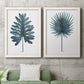 Set of two white barnwood framed canvas prints depicting a coordinated art pair of large blue-green tropical leaves rendered as modern botanical illustrations above a sofa in a modern living room.