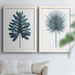 Set of two white barnwood framed canvas prints depicting fan palms, broad philodendron leaves, and crisp white grounds arranged across two complementary compositions above a sofa in a bright living space.