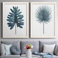 Set of two white barnwood framed canvas prints depicting paired modern botanical illustrations with fan palms, broad philodendron leaves, and crisp white grounds above a sofa in a contemporary living room.