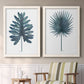 Set of two white barnwood framed canvas prints depicting two modern botanical illustrations featuring large blue-green tropical leaves above a sideboard in a cozy sitting room.