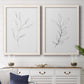 Set of two white barnwood framed canvas prints depicting a coordinated art pair of single continuous-line leafy branches rendered as minimalist botanical line drawings above a dining room sideboard.