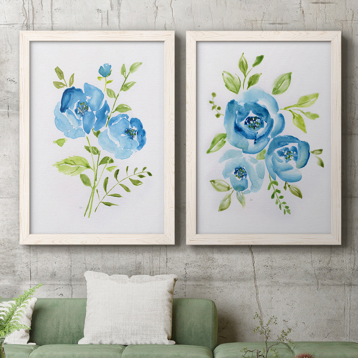 Set of two white barnwood framed canvas prints depicting a coordinated art pair of blue watercolor flowers with fresh green leaves rendered as contemporary botanical paintings above a sofa in a modern living room.
