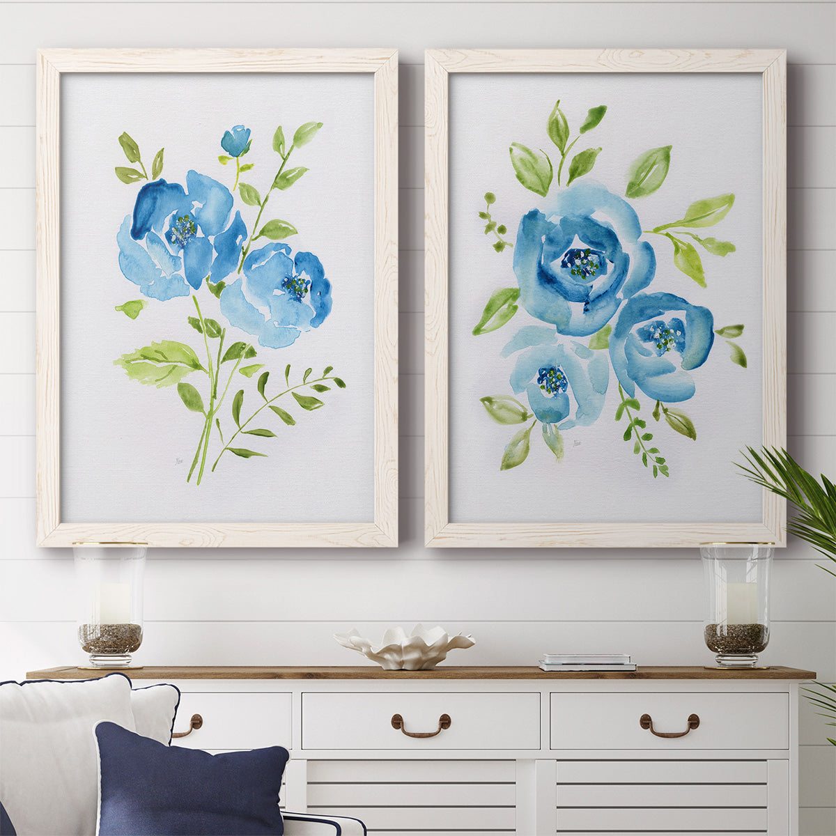 Set of two white barnwood framed canvas prints depicting a coordinated art pair of blue watercolor flowers with fresh green leaves rendered as contemporary botanical paintings above a dining room sideboard.