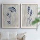 Set of two white barnwood framed canvas prints depicting indigo blossoms, thin stems, and softly distressed gray backgrounds arranged across two complementary compositions above a sofa in a bright living space.