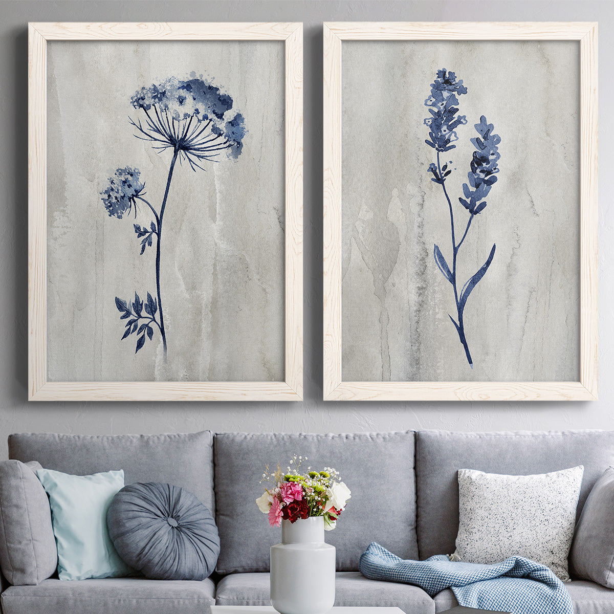 Set of two white barnwood framed canvas prints depicting paired rustic monochrome botanical paintings with indigo blossoms, thin stems, and softly distressed gray backgrounds above a sofa in a contemporary living room.