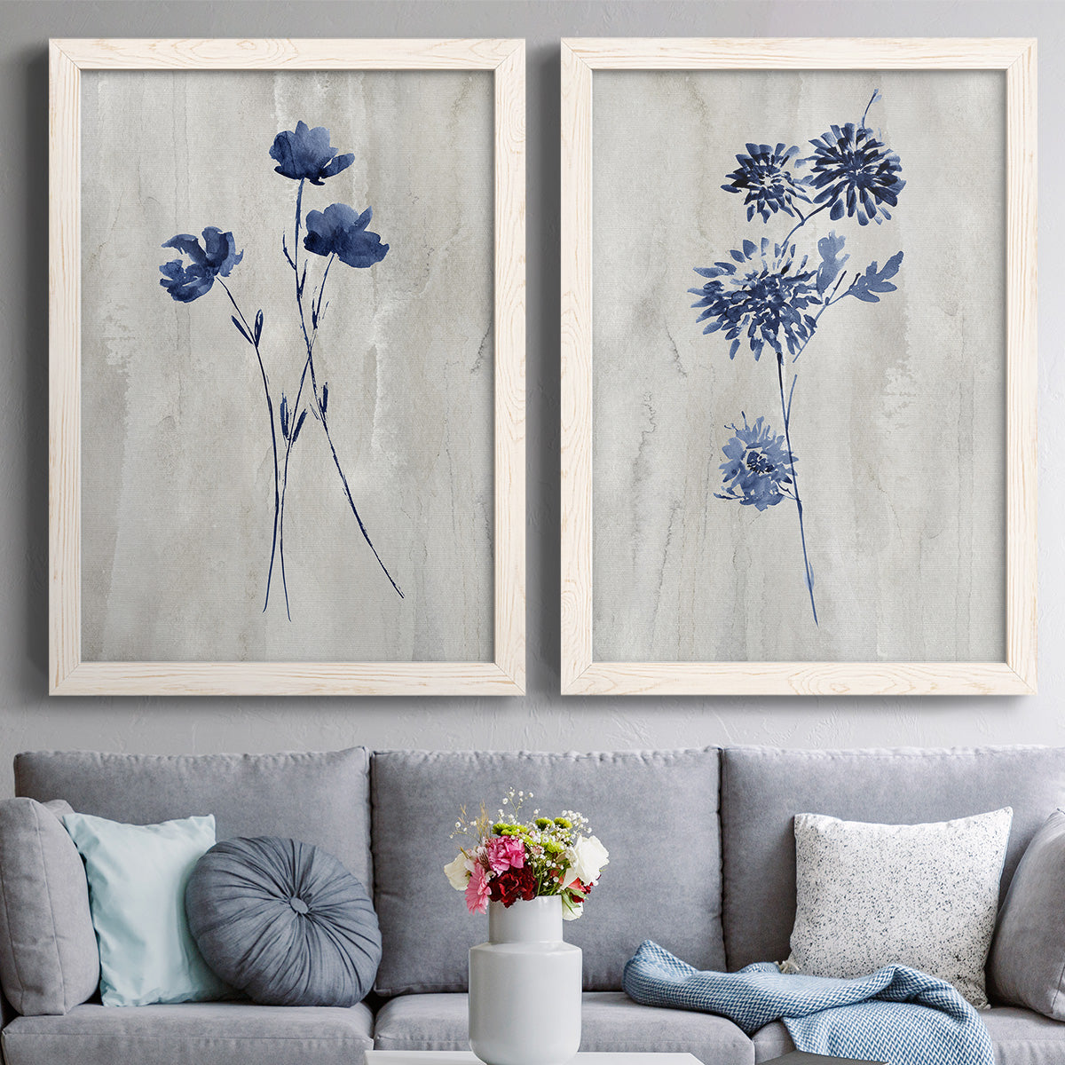 Set of two white barnwood framed canvas prints depicting paired rustic monochrome botanical paintings with indigo blossoms, thin stems, and softly distressed gray backgrounds above a sofa in a contemporary living room.