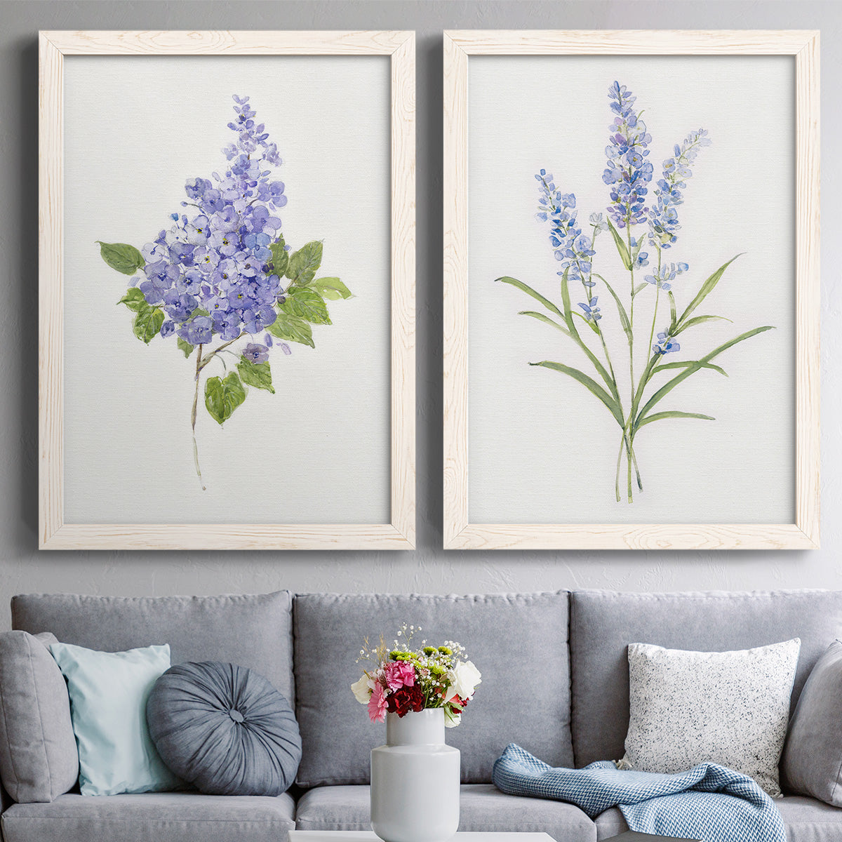Set of two white barnwood framed canvas prints depicting paired delicate watercolor botanical studies with soft blossoms, fine green leaves, and clean white backgrounds above a sofa in a contemporary living room.