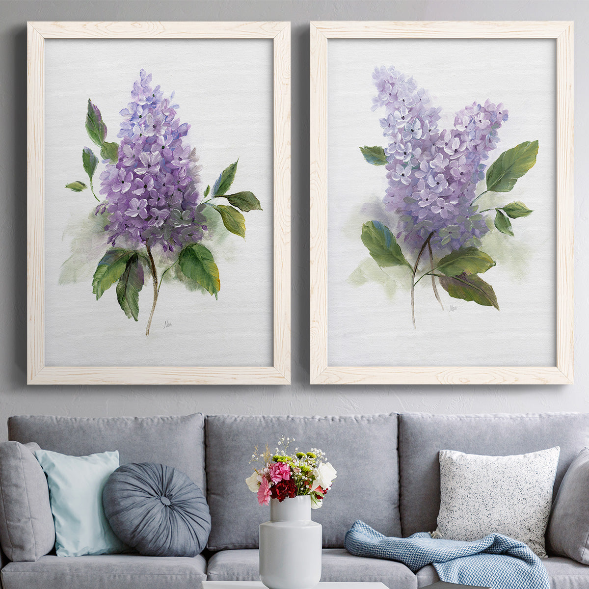 Set of two white barnwood framed canvas prints depicting paired romantic traditional floral paintings with layered lavender blossoms, broad foliage, and softly textured grounds above a sofa in a contemporary living room.