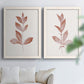 Set of two white barnwood framed canvas prints depicting warm red-brown leaves, slender branches, and soft cream grounds arranged across two complementary compositions above a sofa in a bright living space.