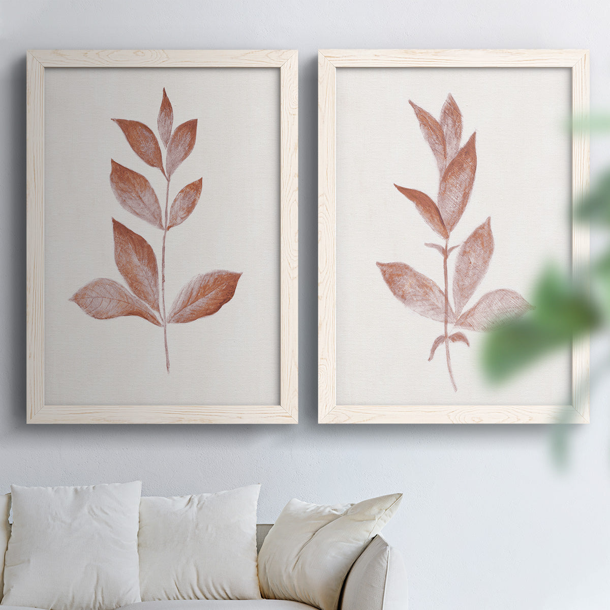 Set of two white barnwood framed canvas prints depicting warm red-brown leaves, slender branches, and soft cream grounds arranged across two complementary compositions above a sofa in a bright living space.