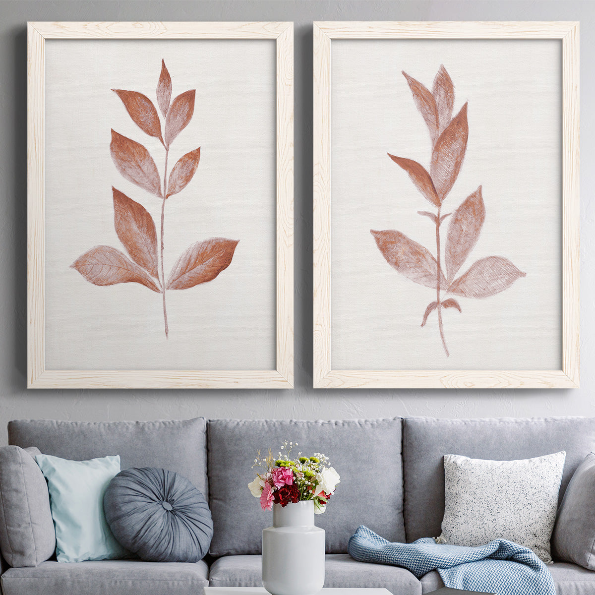 Set of two white barnwood framed canvas prints depicting paired minimalist botanical prints with warm red-brown leaves, slender branches, and soft cream grounds above a sofa in a contemporary living room.