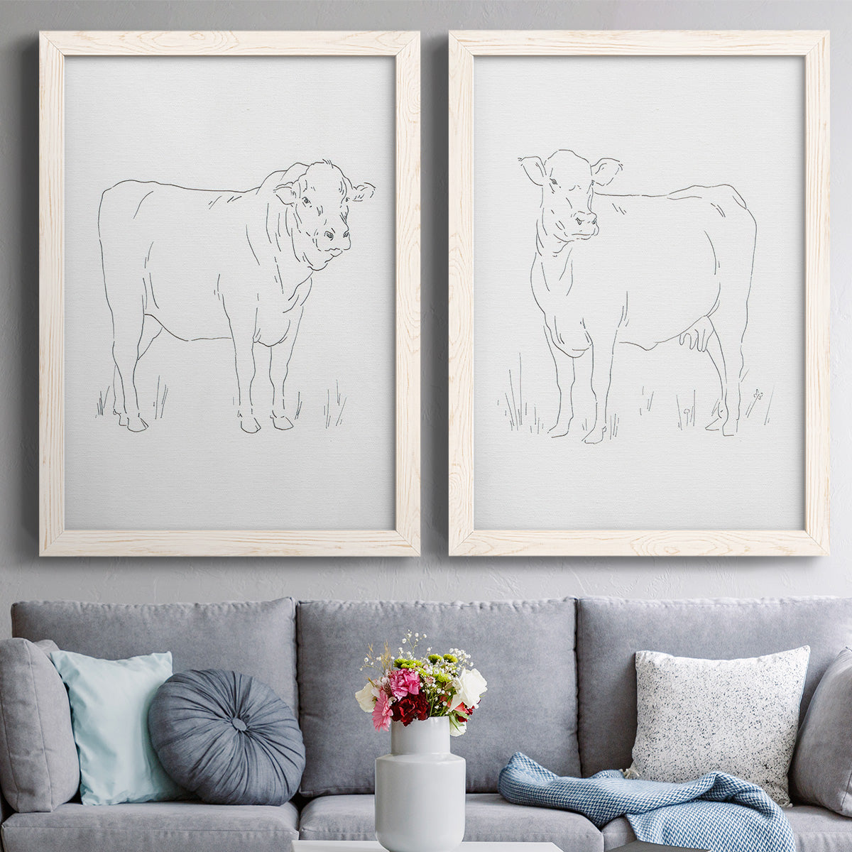 Set of two white barnwood framed canvas prints depicting paired minimalist livestock sketches with fine gray lines, white backgrounds, and quiet farm-animal profiles above a sofa in a contemporary living room.