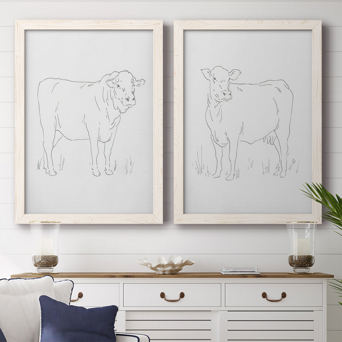 Set of two white barnwood framed canvas prints depicting a coordinated art pair of simple contour drawings of standing cows rendered as minimalist livestock sketches above a dining room sideboard.