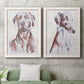 Set of two white barnwood framed canvas prints depicting a coordinated art pair of gentle seated dog portraits rendered as traditional watercolor pet paintings above a sofa in a modern living room.