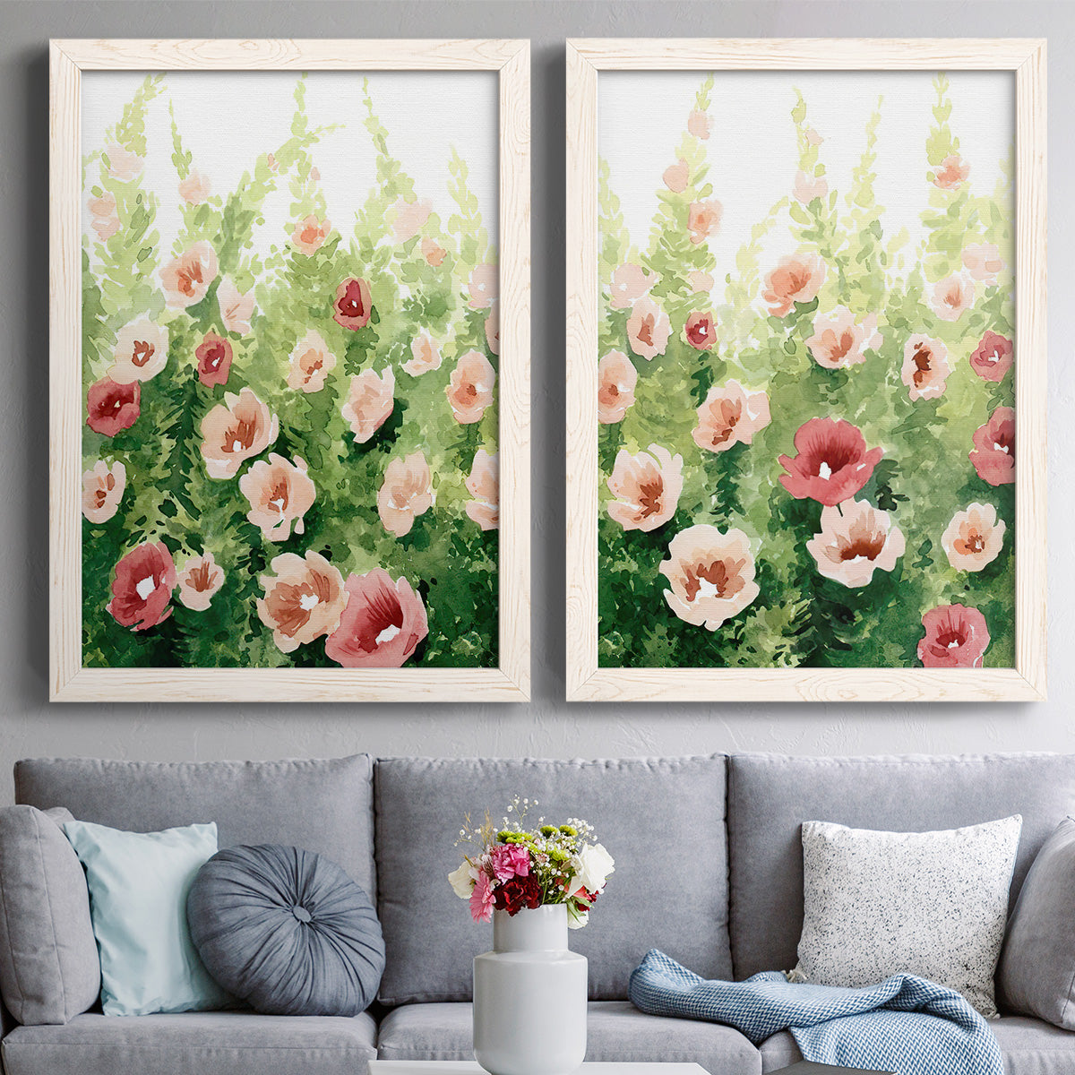Set of two white barnwood framed canvas prints depicting paired luminous impressionistic floral paintings with loose blossoms, green stems, and sunlit watercolor texture above a sofa in a contemporary living room.