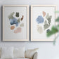 Set of two white barnwood framed canvas prints depicting muted blue green blush and charcoal forms floating on white arranged across two complementary compositions above a sofa in a bright living space.