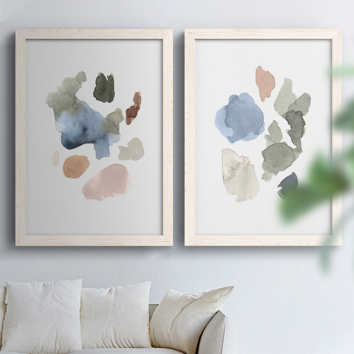 Set of two white barnwood framed canvas prints depicting muted blue green blush and charcoal forms floating on white arranged across two complementary compositions above a sofa in a bright living space.