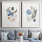 Set of two white barnwood framed canvas prints depicting paired airy minimalist abstracts with muted blue green blush and charcoal forms floating on white above a sofa in a contemporary living room.