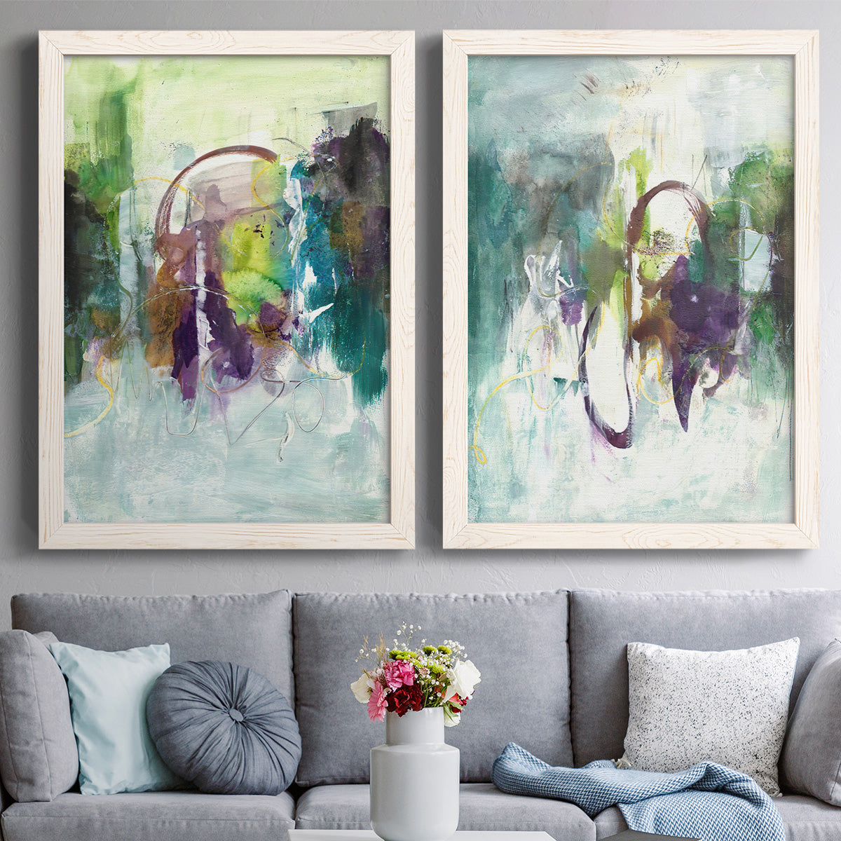 Set of two white barnwood framed canvas prints depicting paired expressive contemporary abstracts with soft-edged color masses, looping lines, and pale blue grounds above a sofa in a contemporary living room.