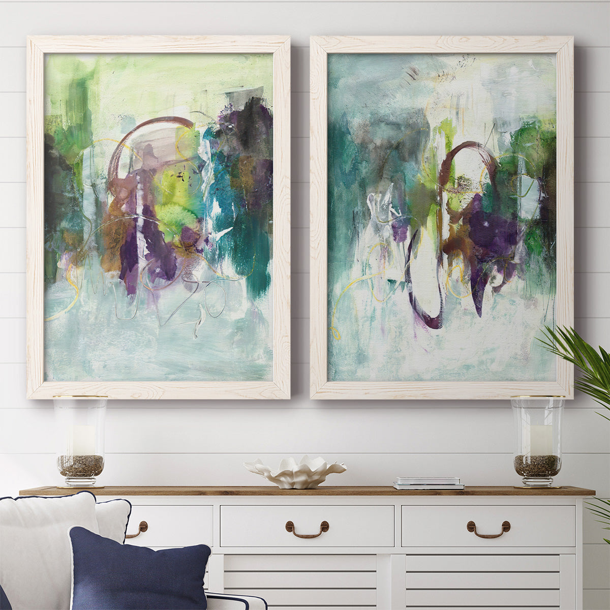 Set of two white barnwood framed canvas prints depicting a coordinated art pair of layered green purple and gray painterly forms rendered as expressive contemporary abstracts above a dining room sideboard.