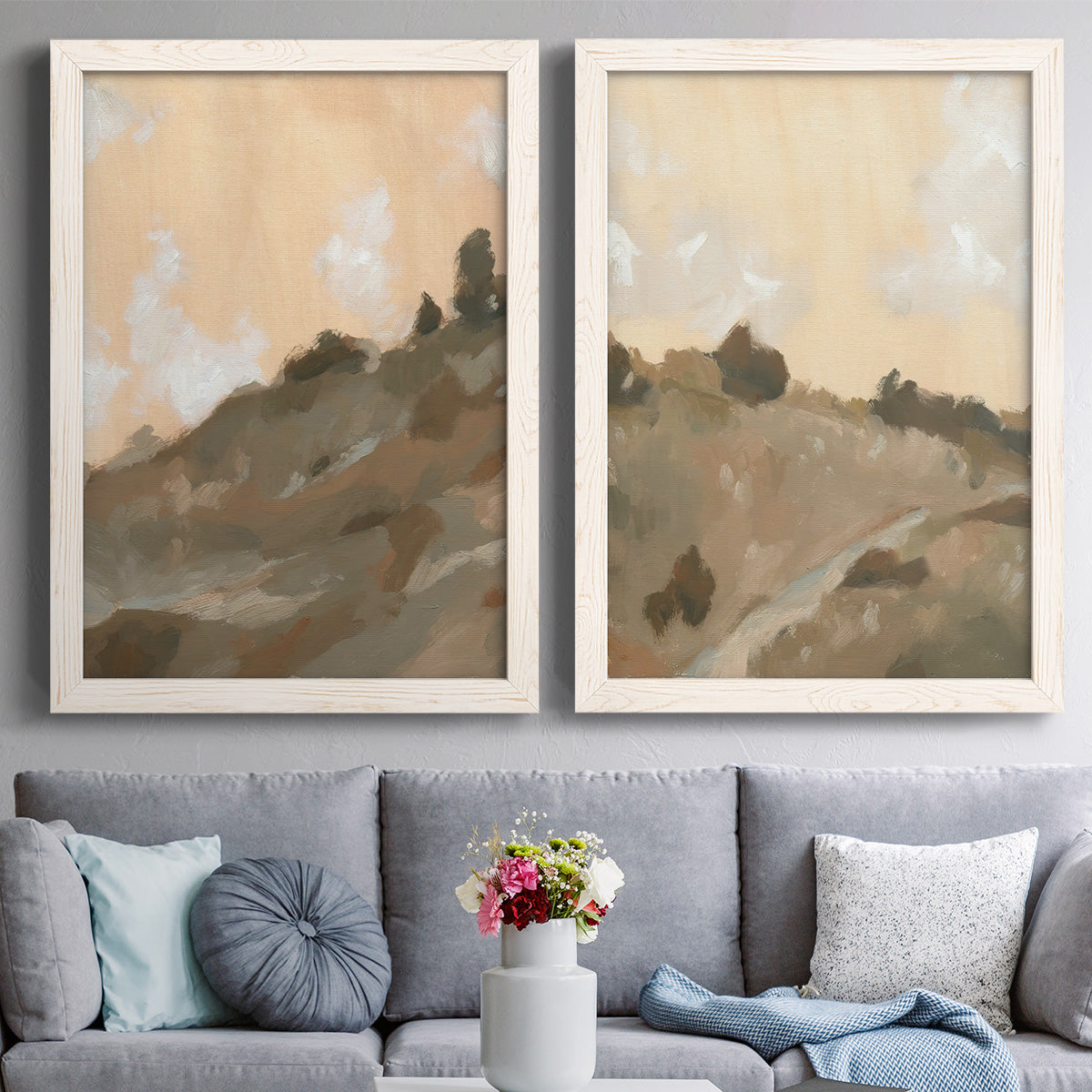 Set of two white barnwood framed canvas prints depicting paired minimalist watercolor landscapes with layered ridges, soft earth tones, and hazy cream skies above a sofa in a contemporary living room.